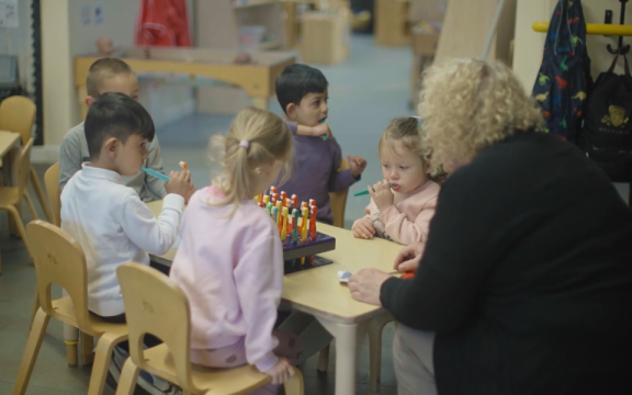 Five children sat at a table in a nursery setting brushing their teeth supported by an adult