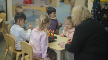 Five children sat at a table in a nursery setting brushing their teeth supported by an adult